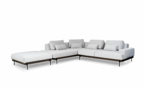 Vista diagonal de la Sala En L Modular Maia con puff, de la marca Aristas. Un diseño contemporáneo, tapizado en tela suave de color neutro, con una base en madera color Chocolate y patas metálicas.