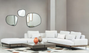 Vista diagonal de la Sala En L Modular Maia con puff, de la marca Aristas. El sofá modular presenta un diseño contemporáneo, con un tapizado en tela suave de color neutro y líneas rectas que aportan elegancia. El puff, con el mismo estilo y material, complementa el conjunto.