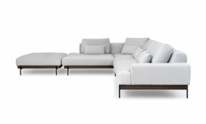 Vista lateral de la Sala En L Modular Maia con puff, de la marca Aristas. Un diseño contemporáneo, tapizado en tela suave de color neutro, base en madera y patas metálicas. El puff, con el mismo estilo y material, complementa el conjunto.