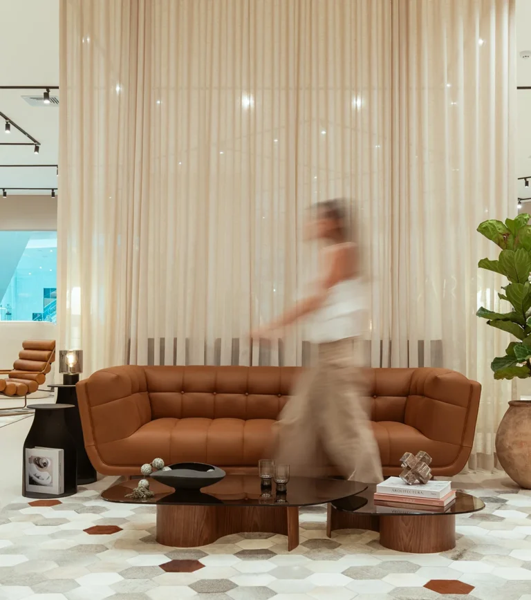 Visitante recorriendo el showroom de Aristas junto a un sofá en cuero cognac, con cortinas translúcidas y mesas de centro en madera de fondo