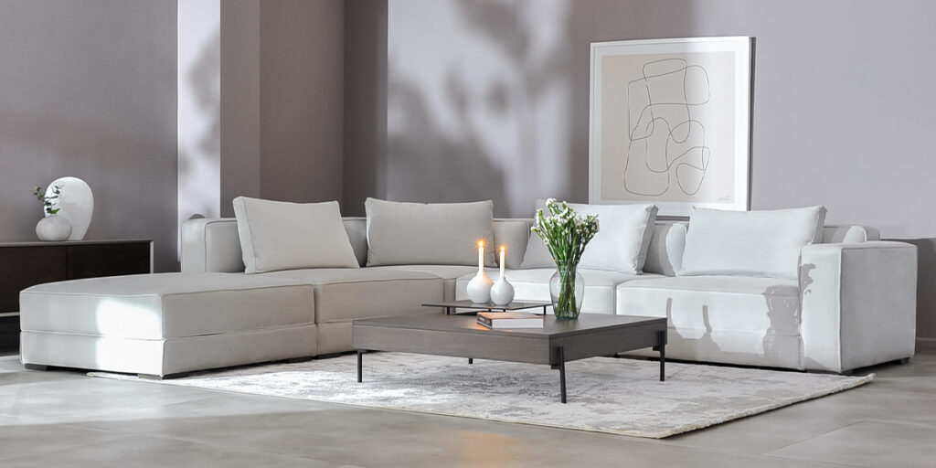 Sofá modular en L en tela clara con mesa de centro gris, velas y flores, bajo un cuadro de línea abstracta