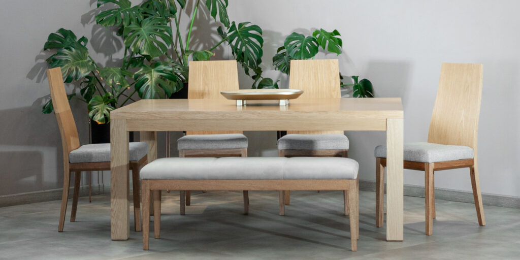 Comedor con mesa de madera clara, sillas y banca con asiento gris, frente a plantas de hojas grandes