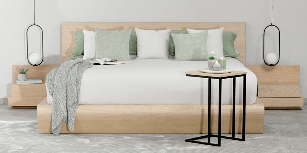 Cama de madera clara con ropa blanca, cojines verde salvia y manta, con lámparas colgantes y una mesa auxiliar negra