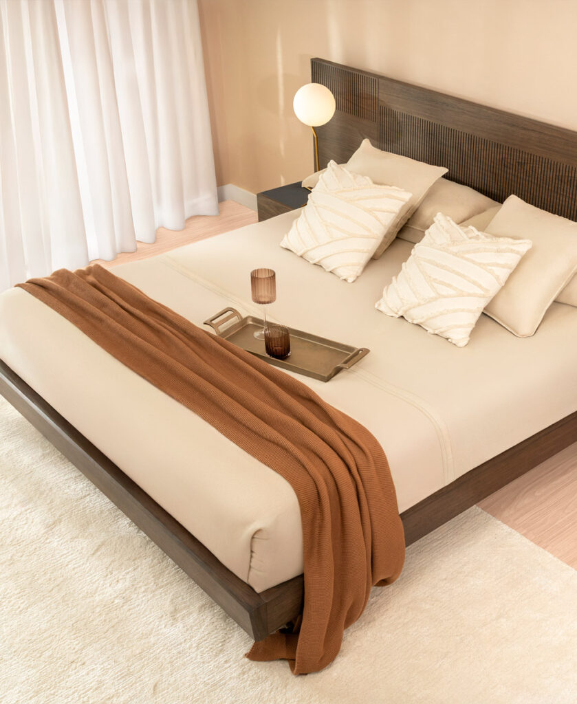 Cama con espaldar en madera oscura ranurada, ropa de cama en tono crudo y manta café tejida, con bandeja y lámpara esférica