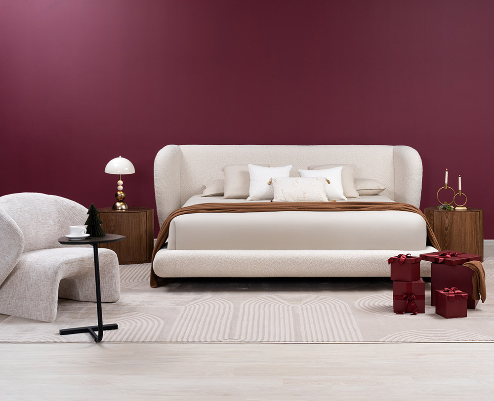 Cama con espaldar alto en bouclé crudo sobre muro vino tinto, con manta terracota, mesas de noche en nogal y regalos