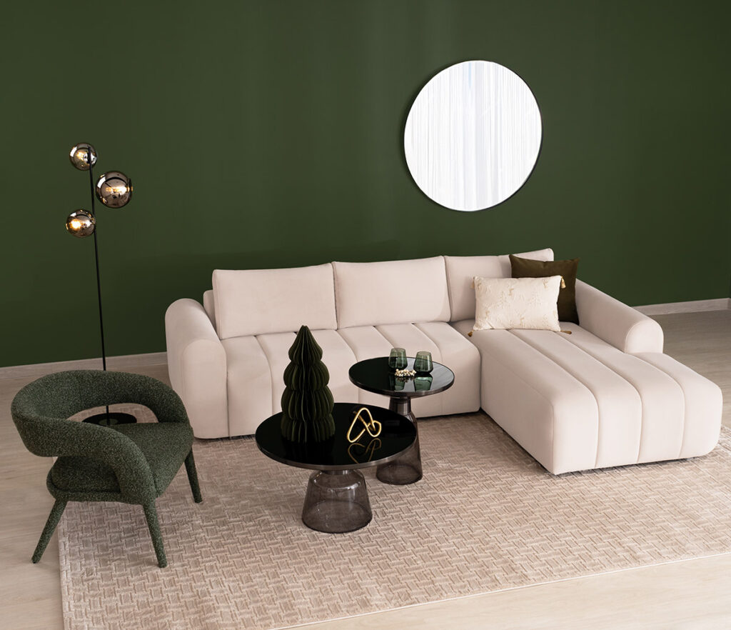 Sala con sofá en L color crudo y chaise longue sobre muro verde, con poltrona en bouclé verde y mesas de centro negras