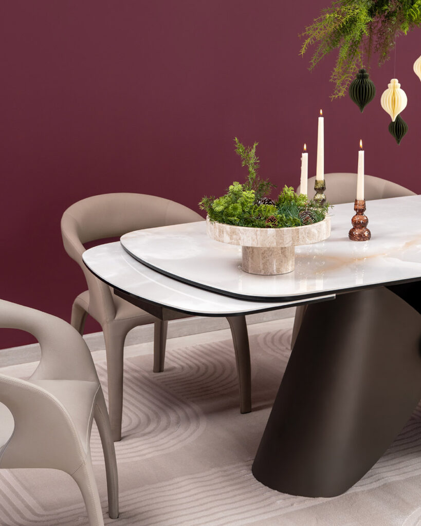 Mesa de comedor en piedra sobre muro vino tinto, con sillas en cuero tono arena, centro de follaje y velas, y adornos colgantes