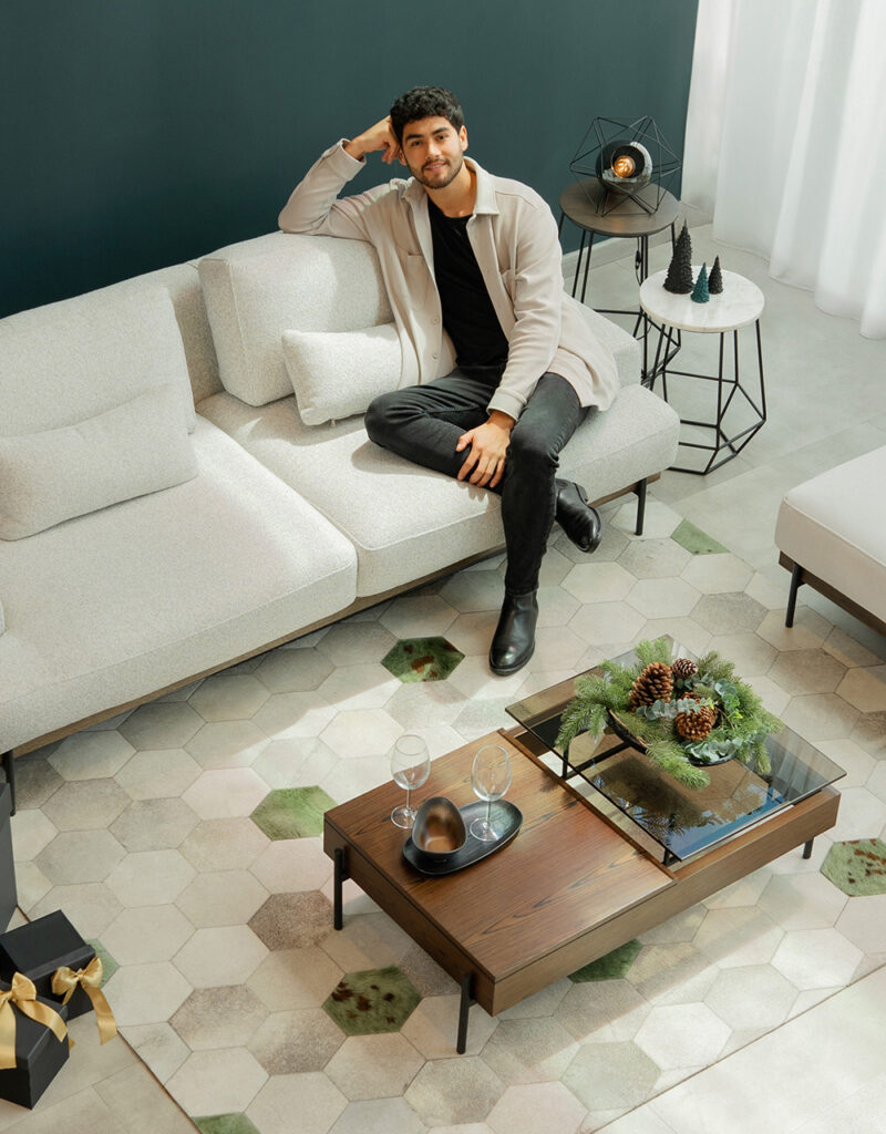 Vista cenital de un hombre sentado en un modular blanco, con mesa de centro de madera y vidrio, copas y un centro de mesa de pino y piñas, sobre alfombra hexagonal
