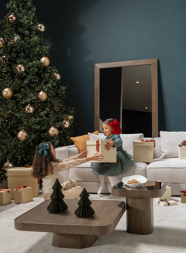 Dos niñas intercambiando un regalo frente a un árbol de Navidad, con sofá blanco, espejo de marco de madera y mesas de centro de madera