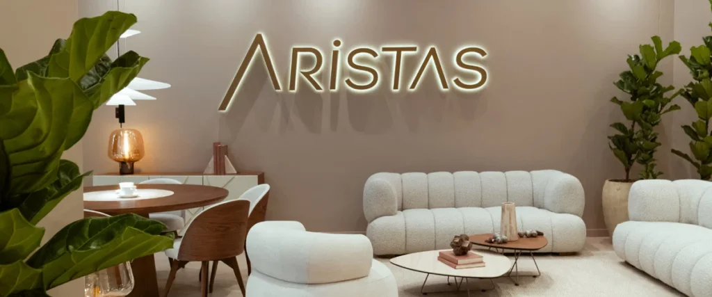 Logo iluminado de Aristas en el muro del showroom, con sofás en bouclé blanco y un comedor redondo en madera