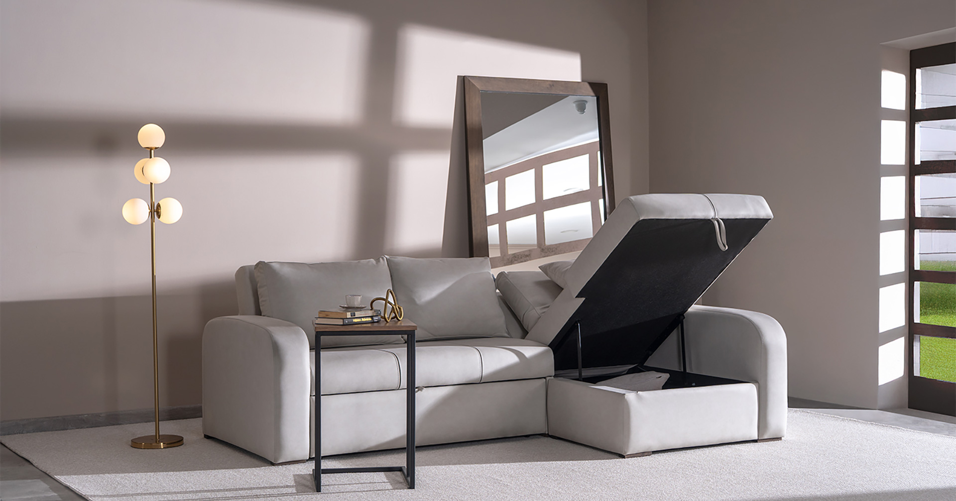 Sofá cama en cuero gris con chaise de almacenamiento abierta, junto a un espejo de piso y una lámpara en latón