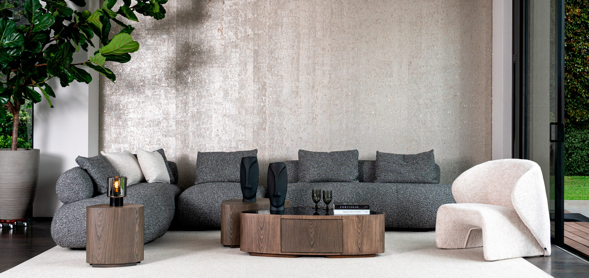Sala con sofá modular en tela gris jaspeada frente a un muro texturizado, mesa de centro redonda en madera y poltrona en tono crudo