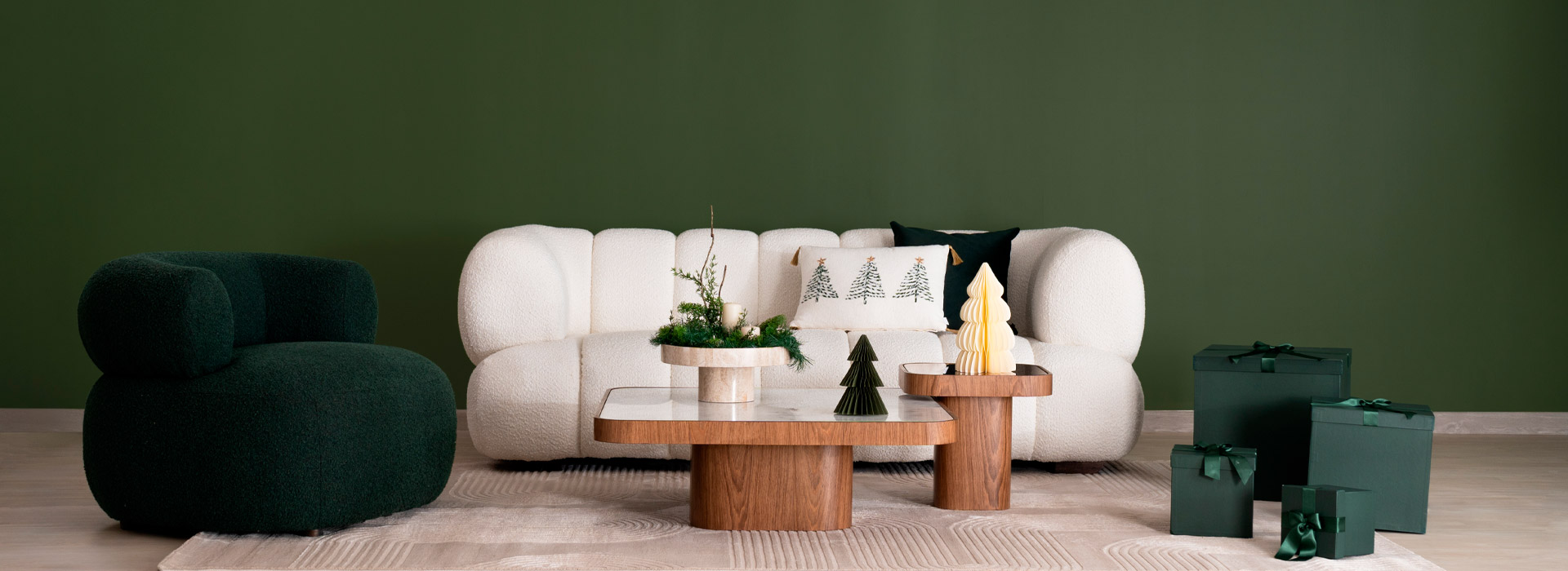 Sofá en bouclé blanco de volúmenes redondeados sobre muro verde, con poltrona verde oscuro, mesas en madera y regalos