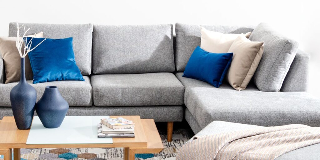 Sofá modular en L gris con cojines azules y beige, y mesa de centro de madera con libros y jarrones azul oscuro