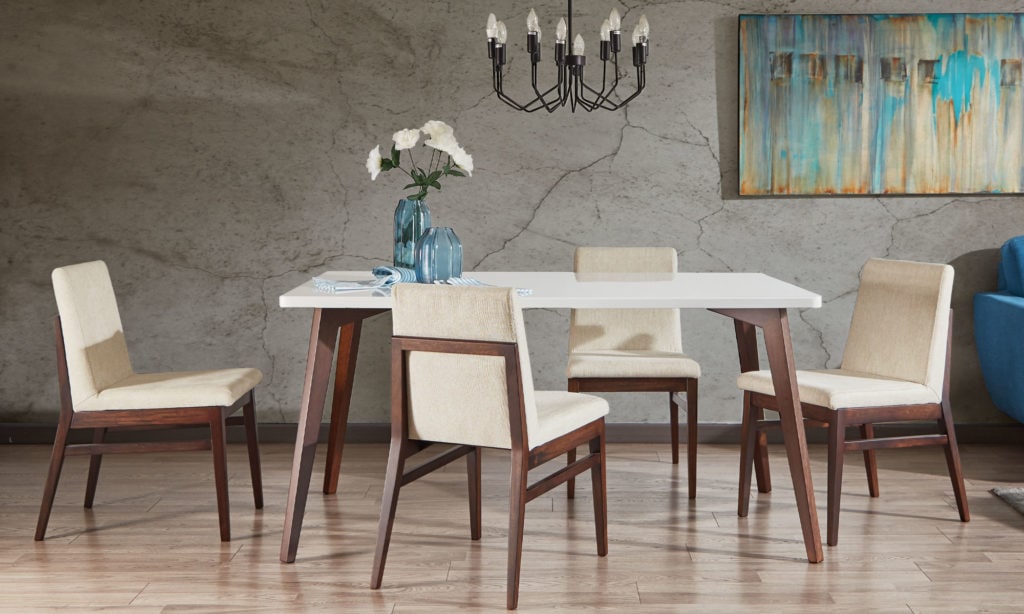 Comedor con mesa de tapa blanca y sillas de madera con asiento crema, bajo una lámpara de brazos frente a un muro de concreto con un cuadro abstracto
