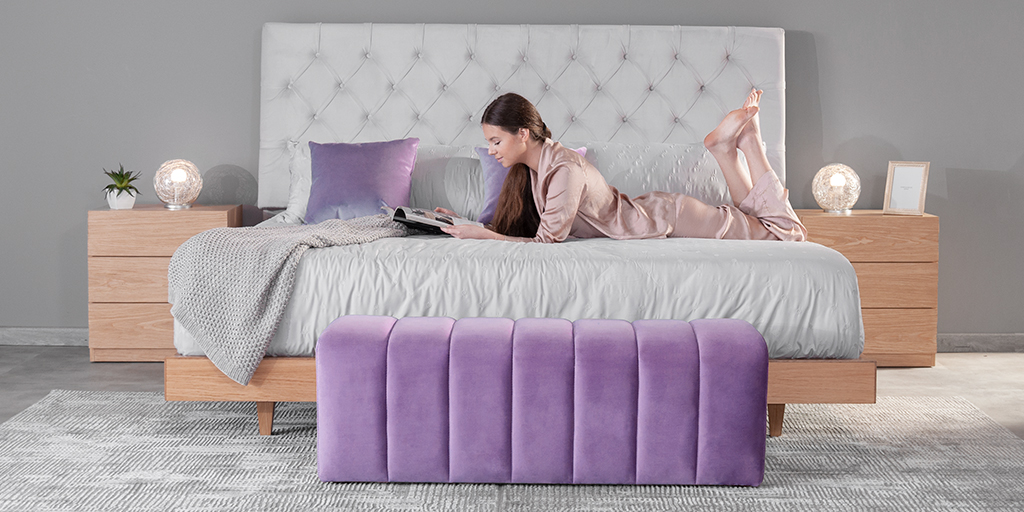Mujer leyendo una revista sobre una cama con cabecero capitoné gris, cojín lila y mesas de noche en madera, con un banco tapizado en lila al pie