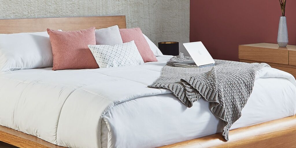 Cama con ropa blanca, cojines rosa y una manta gris tejida, con un computador portátil encima