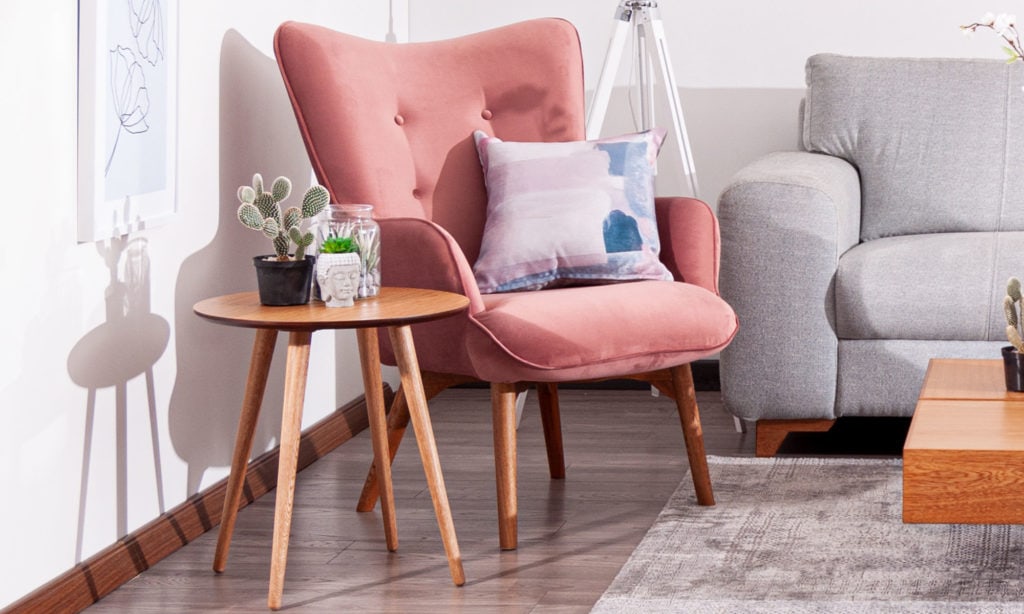 Butaca rosa de respaldo alto con cojín estampado y mesa auxiliar redonda de madera, junto a un sofá gris