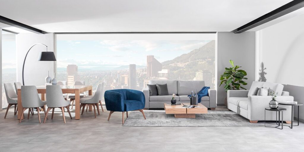 Sala y comedor amplios con sofá gris, poltrona azul y comedor de madera clara, frente a un gran ventanal con vista a una ciudad y montañas