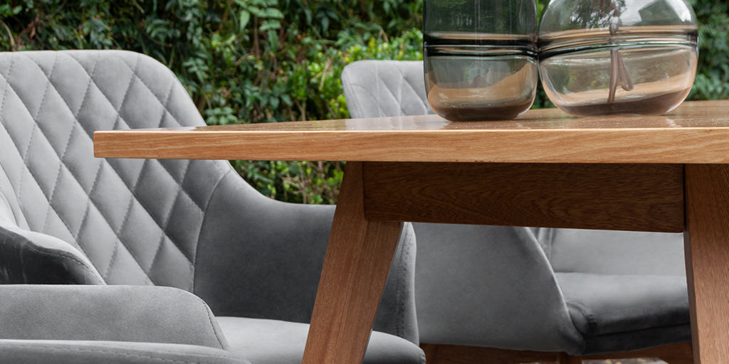 Detalle de una mesa de comedor de madera clara y sillas tapizadas en gris con capitoné de rombos, junto a dos jarrones de vidrio