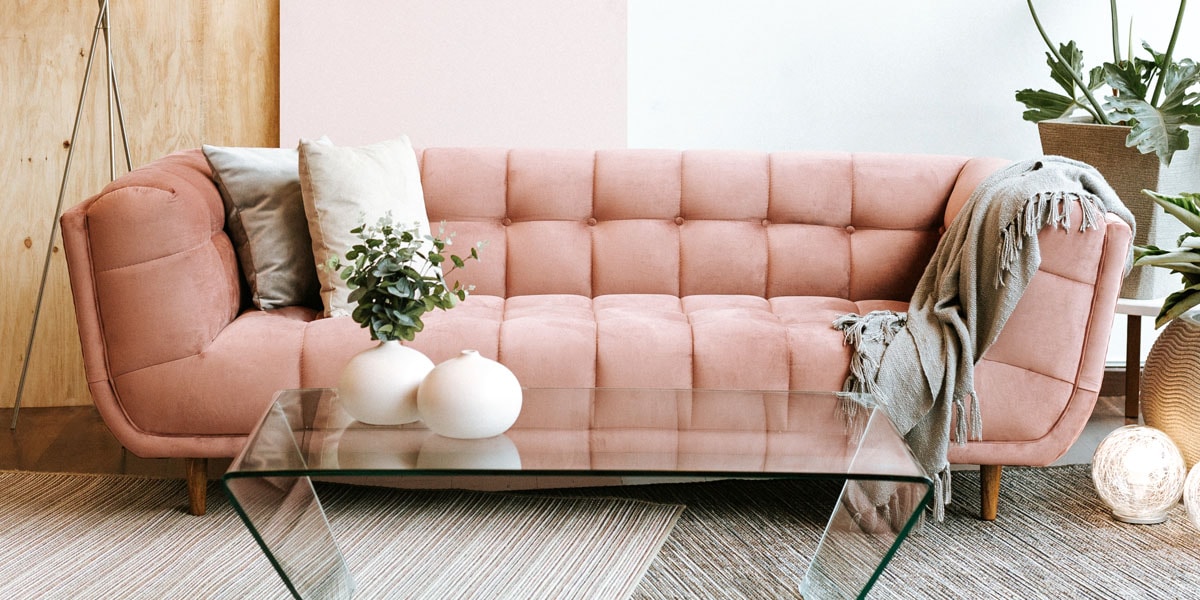 Sofá rosa palo capitoné con una manta y una mesa de centro de vidrio, junto a una planta de hojas grandes