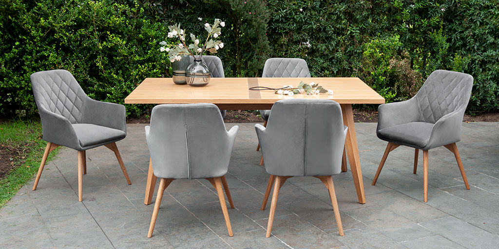 Comedor al aire libre con mesa de madera clara y seis sillas tapizadas en gris con patas de madera, rodeado de vegetación