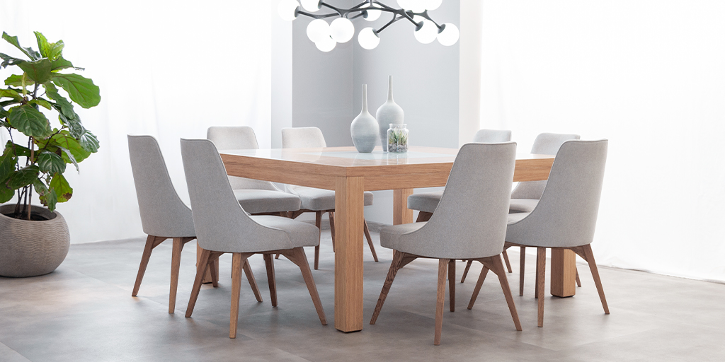 Comedor con mesa de madera clara y seis sillas tapizadas en gris, con una planta de hojas grandes y lámparas blancas colgantes