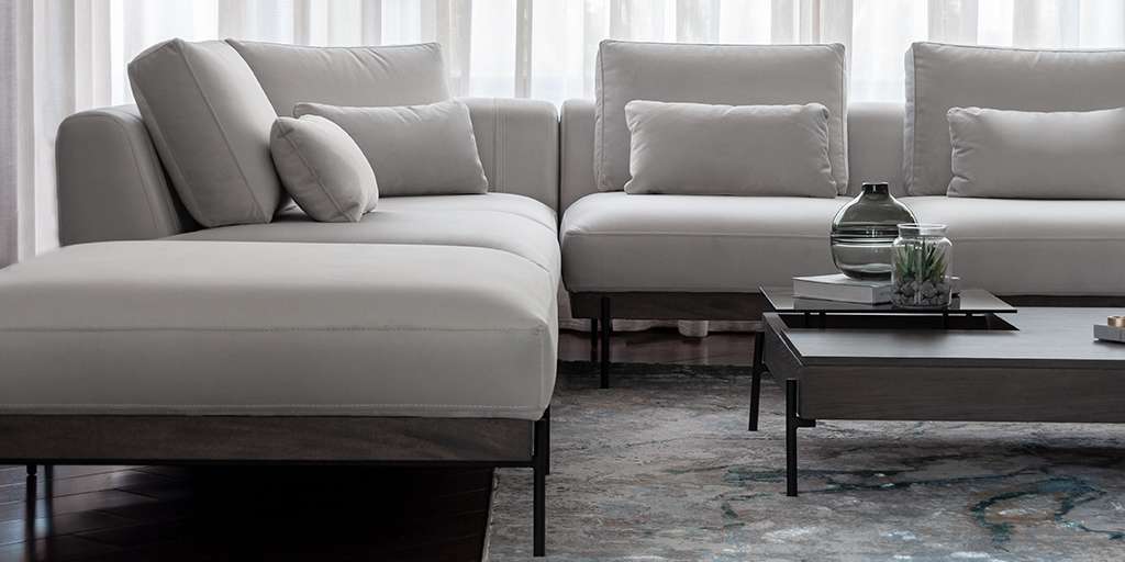 Sofá modular en L tapizado en gris claro con cojines y mesa de centro oscura, frente a un ventanal con cortinas blancas
