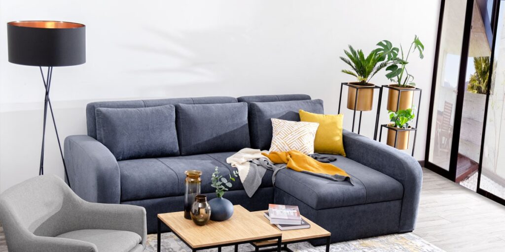 Sofá cama gris azulado con chaise longue, cojines amarillo y estampado, una manta, mesas de centro de madera, una butaca gris y plantas en soportes dorados