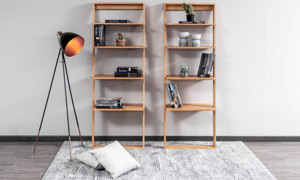 Dos bibliotecas de madera clara con libros y objetos y una lámpara de trípode negra, junto a cojines blancos en el piso