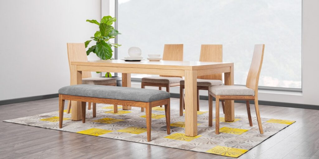 Comedor de madera clara con banca y sillas de asiento gris, sobre un tapete con detalles amarillos frente a un ventanal