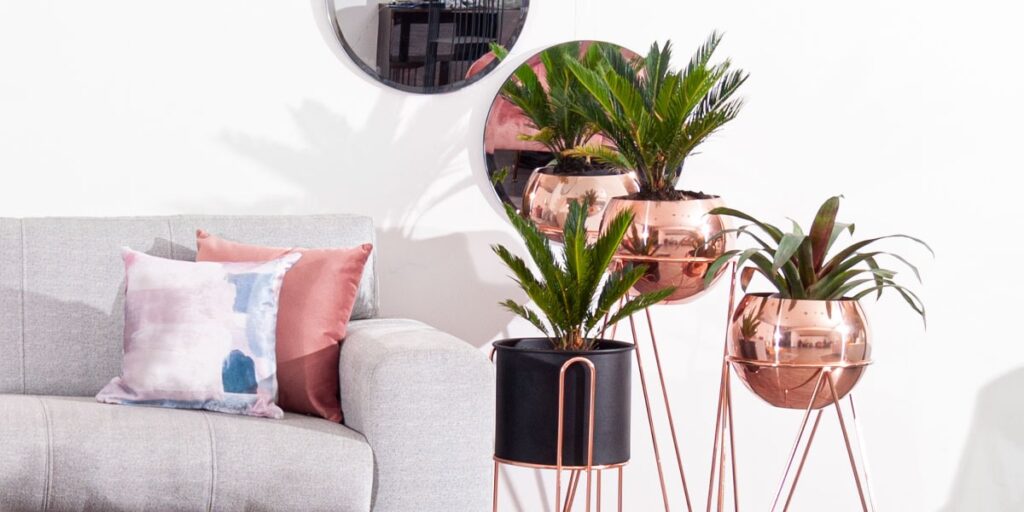 Sofá gris con cojines rosa y estampados, junto a plantas en soportes de cobre y un espejo redondo