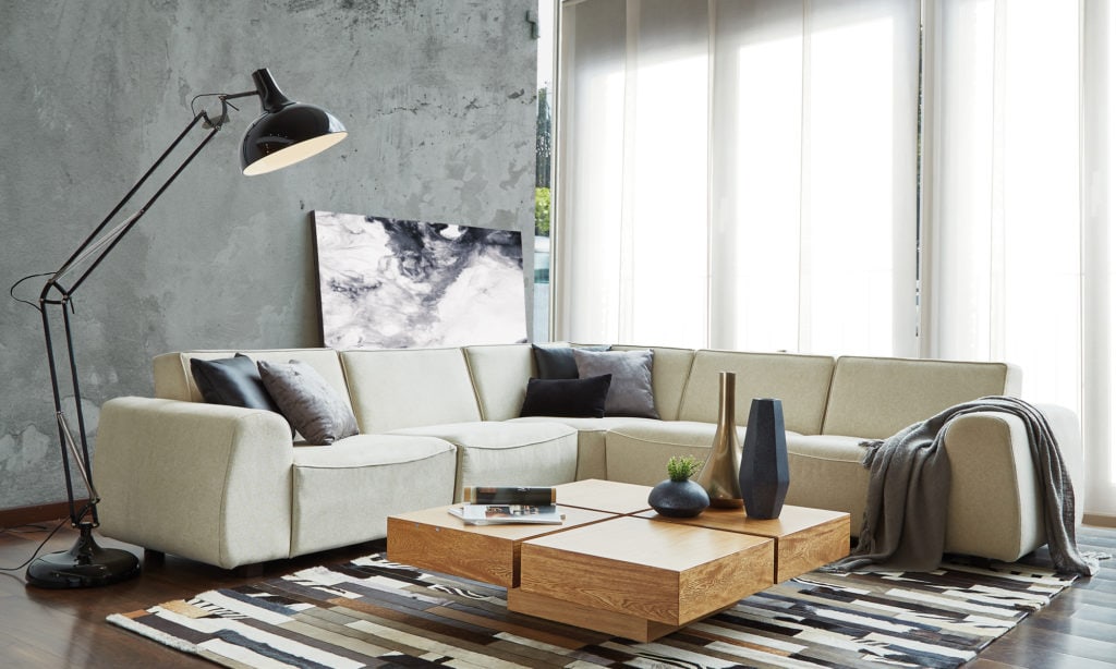 Sofá modular crema con cojines y manta gris, mesa de centro de madera de cuatro módulos y una lámpara de arco, sobre un tapete de rayas