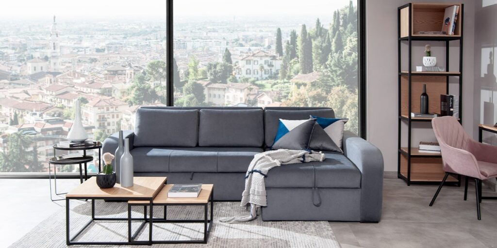 Sofá cama gris azulado con chaise longue, mesas de centro de madera y una silla rosa, frente a un ventanal con vista a una ciudad
