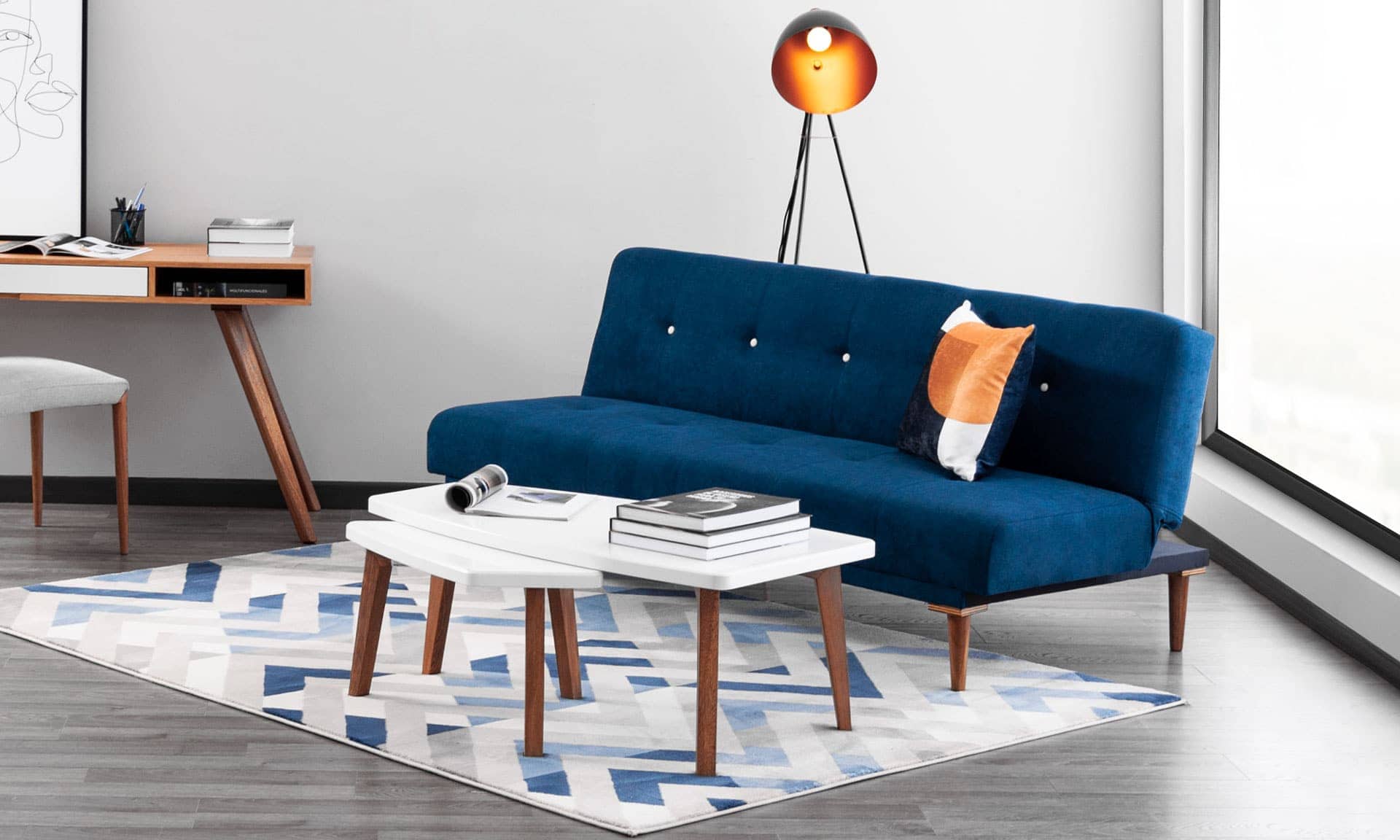 Sofá cama azul oscuro con cojín naranja y dos mesas de centro blancas, junto a un escritorio y una lámpara de trípode