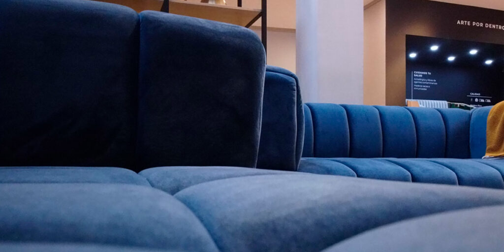 Detalle de un sofá azul de canales verticales en un showroom, con una pantalla al fondo