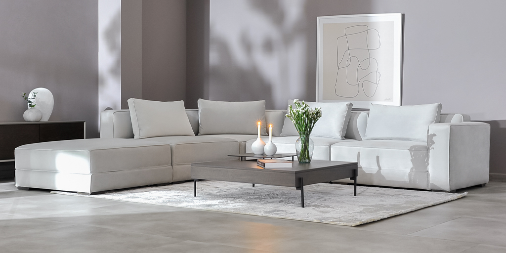 Sala con sofá modular en L en tela blanca, mesa de centro gris, flores y velas, bajo un cuadro de línea abstracta
