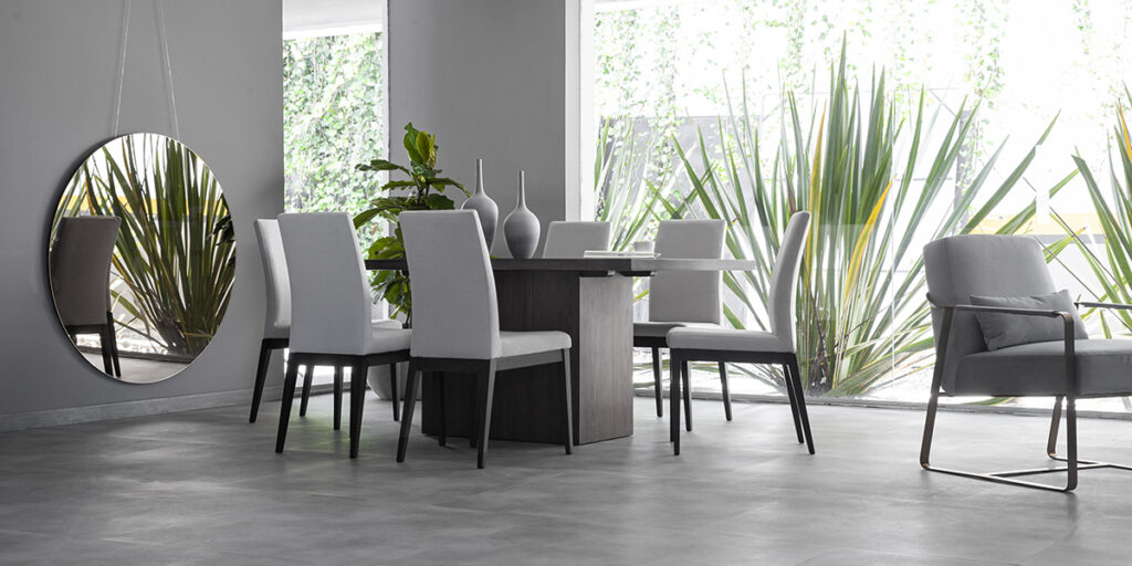 Comedor con mesa oscura de base cilíndrica y sillas grises claras, junto a un espejo redondo y una poltrona de metal