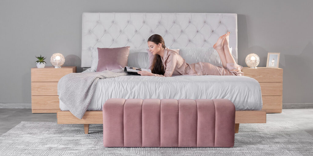 Mujer recostada leyendo una revista sobre una cama con cabecero capitoné blanco y un banco rosa al pie, entre mesas de noche de madera