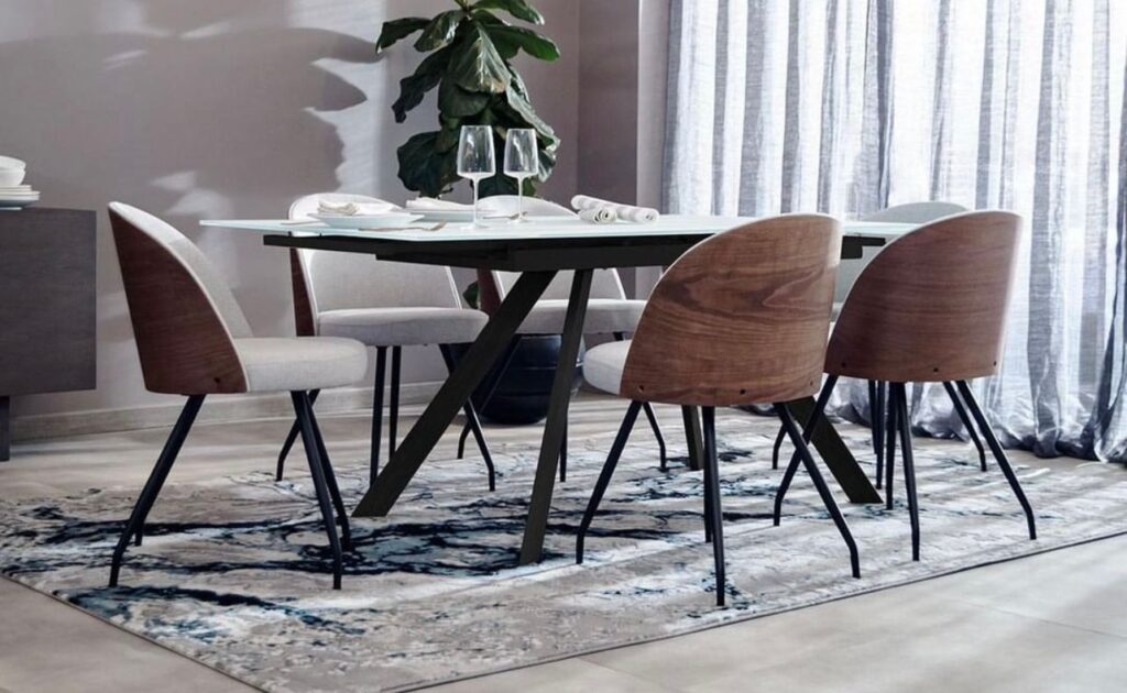 Comedor con mesa de tapa clara y patas negras en X, y sillas con respaldo de madera de nogal y asiento gris, sobre un tapete con vetas azules