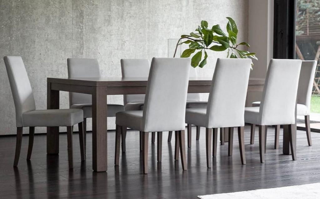 Comedor largo con mesa de madera oscura y ocho sillas tapizadas en gris claro frente a un muro de concreto, con una planta