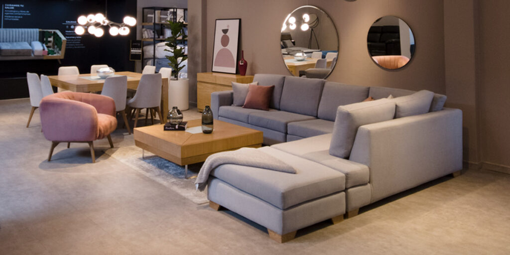 Showroom con sofá modular en L gris, poltrona rosa, mesa de centro de madera y un comedor con espejos redondos en el muro