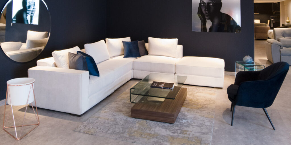 Sofá modular blanco y poltrona azul oscuro con mesa de centro de vidrio frente a un muro azul oscuro con cuadros, en un showroom