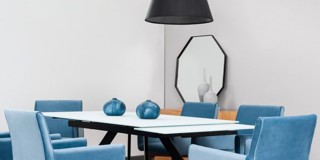 Comedor con mesa de tapa blanca, sillas azules y un espejo octagonal de marco negro en el muro