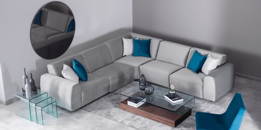 Sala con sofá modular en L gris y cojines azul petróleo, mesa de centro de vidrio y un espejo redondo oscuro en el muro