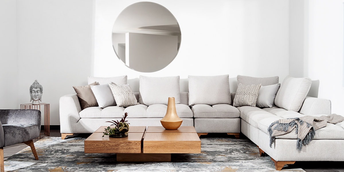 Sala con sofá modular en L crema y mesa de centro de madera, bajo un espejo redondo en el muro, con una poltrona de terciopelo gris
