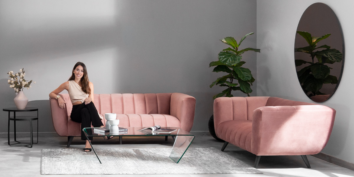 Mujer sentada en un sofá rosa de canales verticales junto a un sillón rosa y una mesa de vidrio, frente a un espejo ovalado y una planta