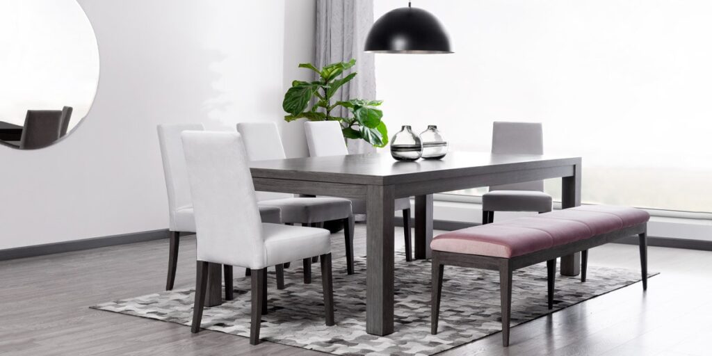 Comedor con mesa oscura, sillas grises claras y banca rosa, bajo una lámpara negra y junto a una planta de hojas grandes