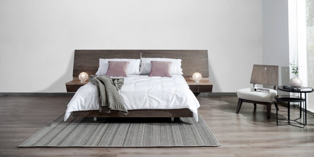Dormitorio con cama de cabecero y base de madera oscura, ropa blanca y cojines rosa, mesas de noche con lámparas y una poltrona blanca