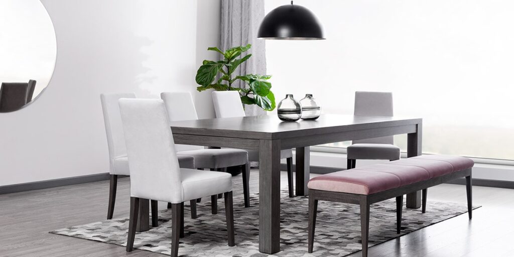 Comedor con mesa oscura, sillas blancas y banca rosa, bajo una lámpara negra y junto a una planta de hojas grandes